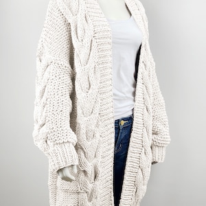 May include: A cream-colored cable knit cardigan with long sleeves and a deep V-neck. The cardigan is made of a chunky yarn and has a relaxed fit.