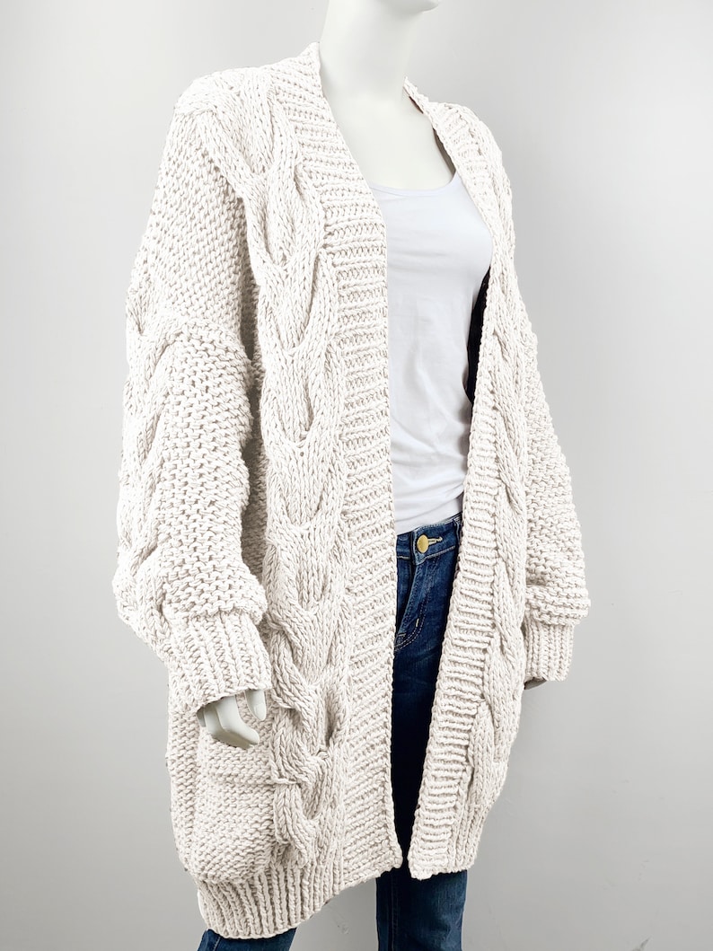 May include: A cream-colored cable knit cardigan with long sleeves and a deep V-neck. The cardigan is made of a chunky yarn and has a relaxed fit.