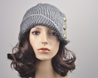 May include: A gray knit hat with a brim and three gold-colored buttons. The hat is made of a thick, textured yarn and has a slight slant to the brim. The hat is displayed on a mannequin head with brown hair.