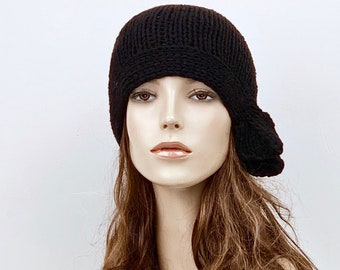 May include: A black knit cloche hat with a brim and a decorative side detail. The hat is made of a thick, textured yarn and has a rounded crown. The hat is displayed on a mannequin with long, wavy brown hair.