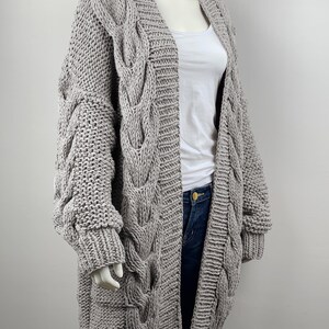 May include: A long, gray cable knit cardigan with a V-neck and long sleeves. The cardigan is made of a chunky yarn and has a relaxed fit.