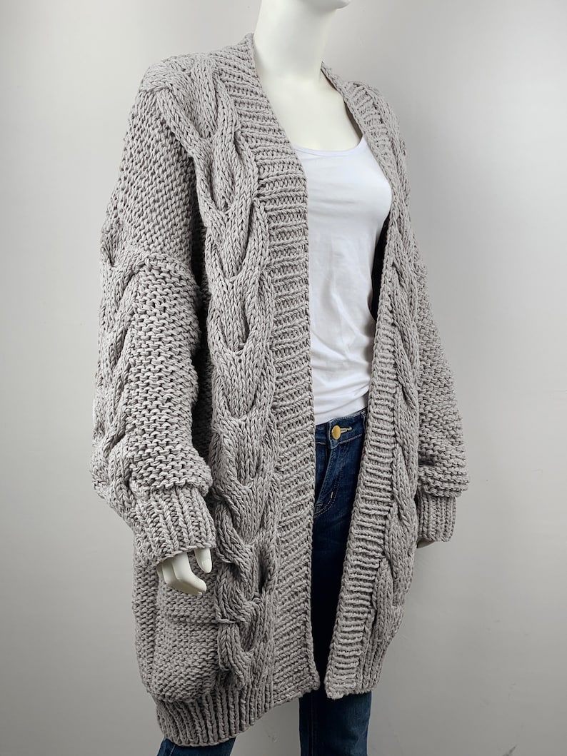May include: A long, gray cable knit cardigan with a V-neck and long sleeves. The cardigan is made of a chunky yarn and has a relaxed fit.