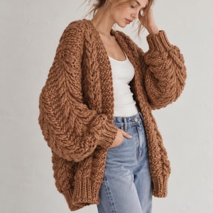 May include: A chunky, oversized, open-front cardigan in a solid brown color, featuring a prominent cable knit pattern on the sleeves and front panels, long sleeves with ribbed cuffs, and a thick ribbed hemline, worn over a white tank top and blue jeans.