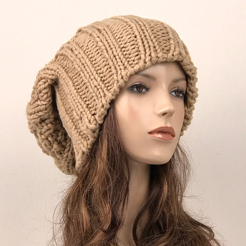 May include: A beige knitted beanie with a slouchy design. The beanie is made of a soft, chunky yarn and has a ribbed texture.