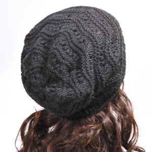 May include: A black knitted beanie with a cable knit pattern. The beanie is worn on a mannequin with long brown curly hair.