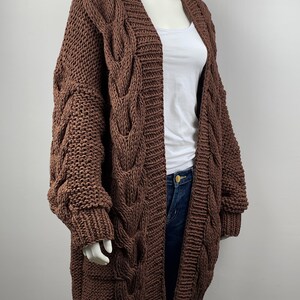 May include: A brown cable knit cardigan with long sleeves and a deep V-neck. The cardigan is made of a chunky yarn and has a relaxed fit.
