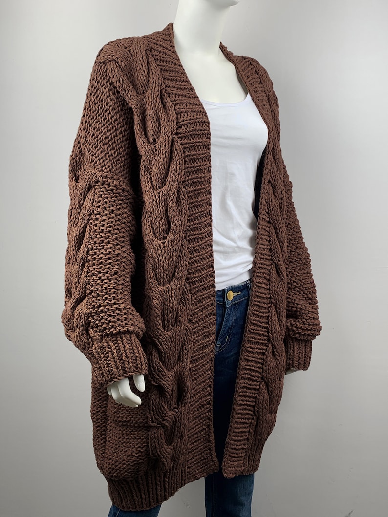 May include: A brown cable knit cardigan with long sleeves and a deep V-neck. The cardigan is made of a chunky yarn and has a relaxed fit.