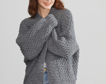 Puede incluir: Un cárdigan de punto gris con un ajuste holgado y una silueta ligeramente oversized. El cárdigan tiene un cuello chal y mangas largas.