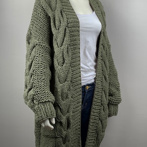 May include: A long, olive green cable knit cardigan with a V-neck and long sleeves. The cardigan is made of a chunky yarn and has a relaxed fit.