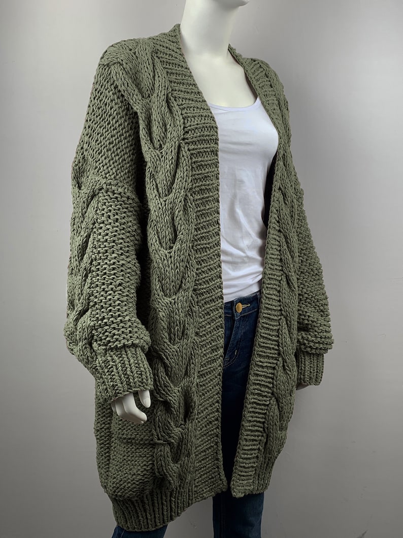 May include: A long, olive green cable knit cardigan with a V-neck and long sleeves. The cardigan is made of a chunky yarn and has a relaxed fit.