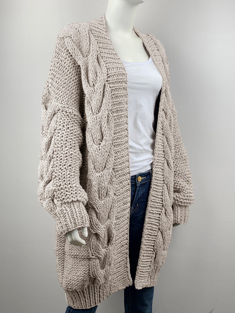 May include: A long, beige cable knit cardigan with a relaxed fit and oversized sleeves. The cardigan is open in the front and has a V-neck.