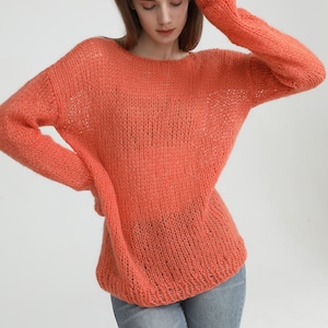 May include: A loose-fitting, coral-colored knitted sweater with a round neckline and long sleeves. The sweater is made of a soft, lightweight yarn and has a slightly open weave.