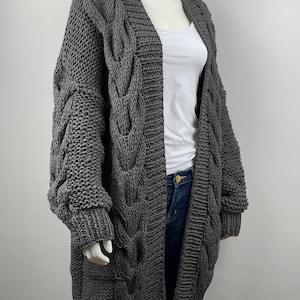 May include: A gray cable knit cardigan with long sleeves and a deep V-neck. The cardigan is made of a chunky yarn and has a relaxed fit.