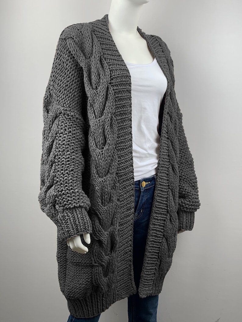 May include: A gray cable knit cardigan with long sleeves and a deep V-neck. The cardigan is made of a chunky yarn and has a relaxed fit.