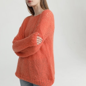 May include: A coral-colored knitted sweater with a loose fit and a slightly open neckline. The sweater is made of a soft, fuzzy yarn and has a relaxed, casual style.