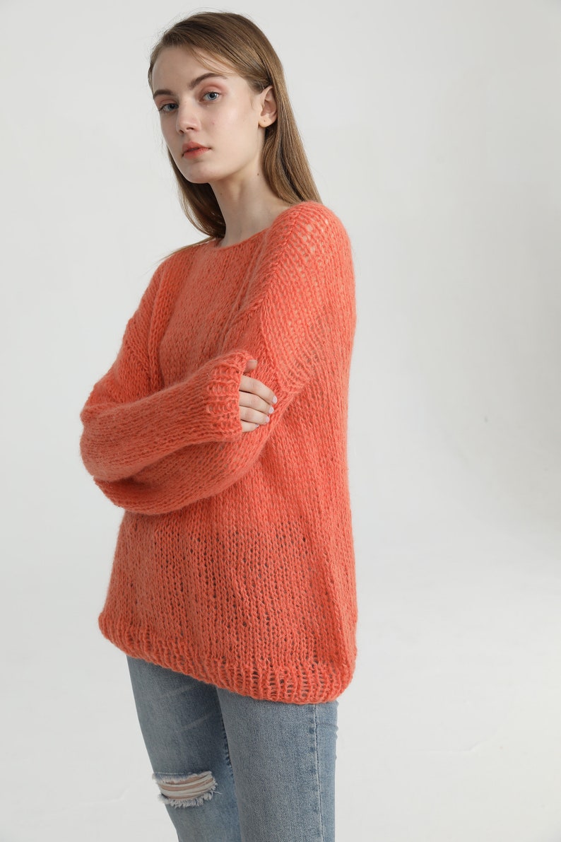 May include: A coral-colored knitted sweater with a loose fit and a slightly open neckline. The sweater is made of a soft, fuzzy yarn and has a relaxed, casual style.