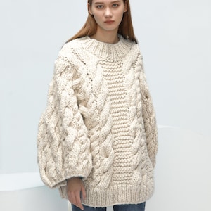 Könnte beinhalten: Cremefarbener Zopfstrickpullover mit hohem Ausschnitt und voluminösen Ärmeln. Der Pullover hat komplizierte Zopfmuster und einen gerippten Saum. Das Model trägt blaue Jeans. Dieser gemütliche Pullover ist perfekt für kaltes Wetter.