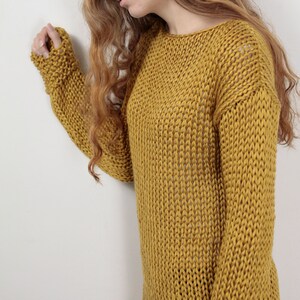 May include: A mustard yellow knitted sweater with a loose fit and a round neckline. The sweater is made of a soft, chunky yarn and has a relaxed, casual style.