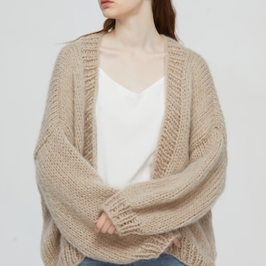 Puede incluir: Un cárdigan de punto beige con cuello en V y mangas largas. El cárdigan está hecho de hilo grueso y tiene un corte holgado.