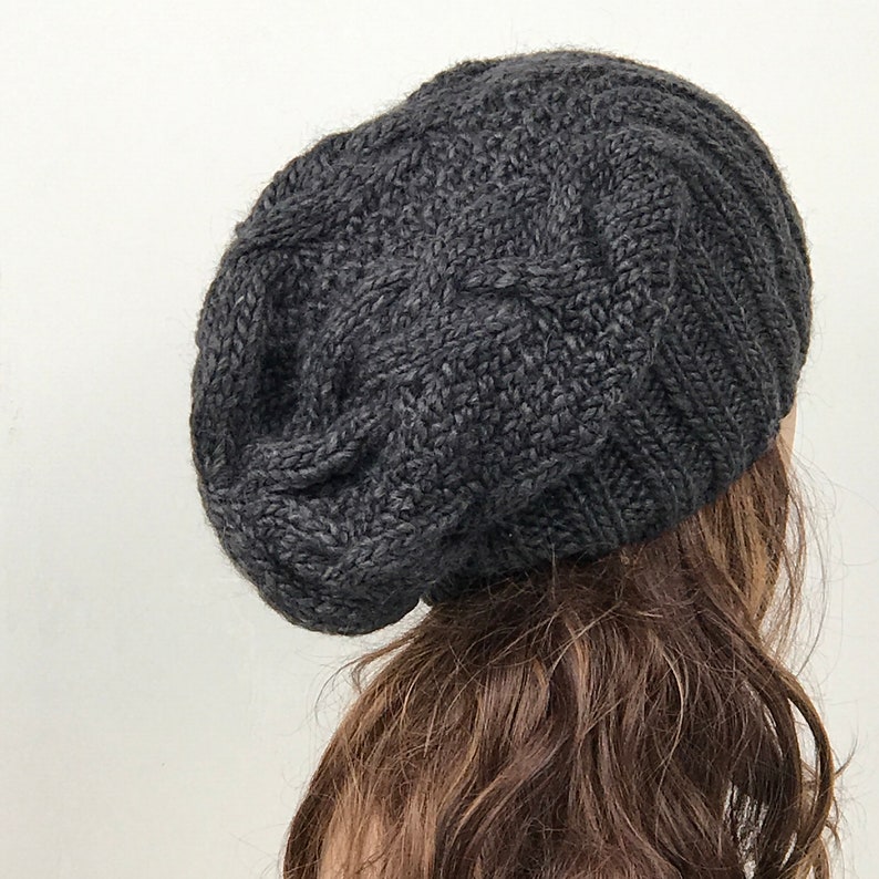 May include: A dark gray knitted beanie with a cable knit pattern. The beanie is slouchy and has a relaxed fit.
