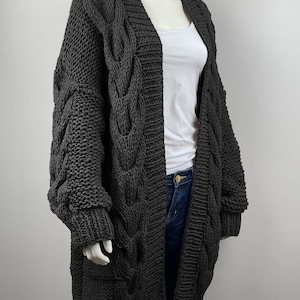 May include: A long, gray cable knit cardigan with a V-neck and long sleeves. The cardigan is made of a chunky yarn and has a relaxed fit.