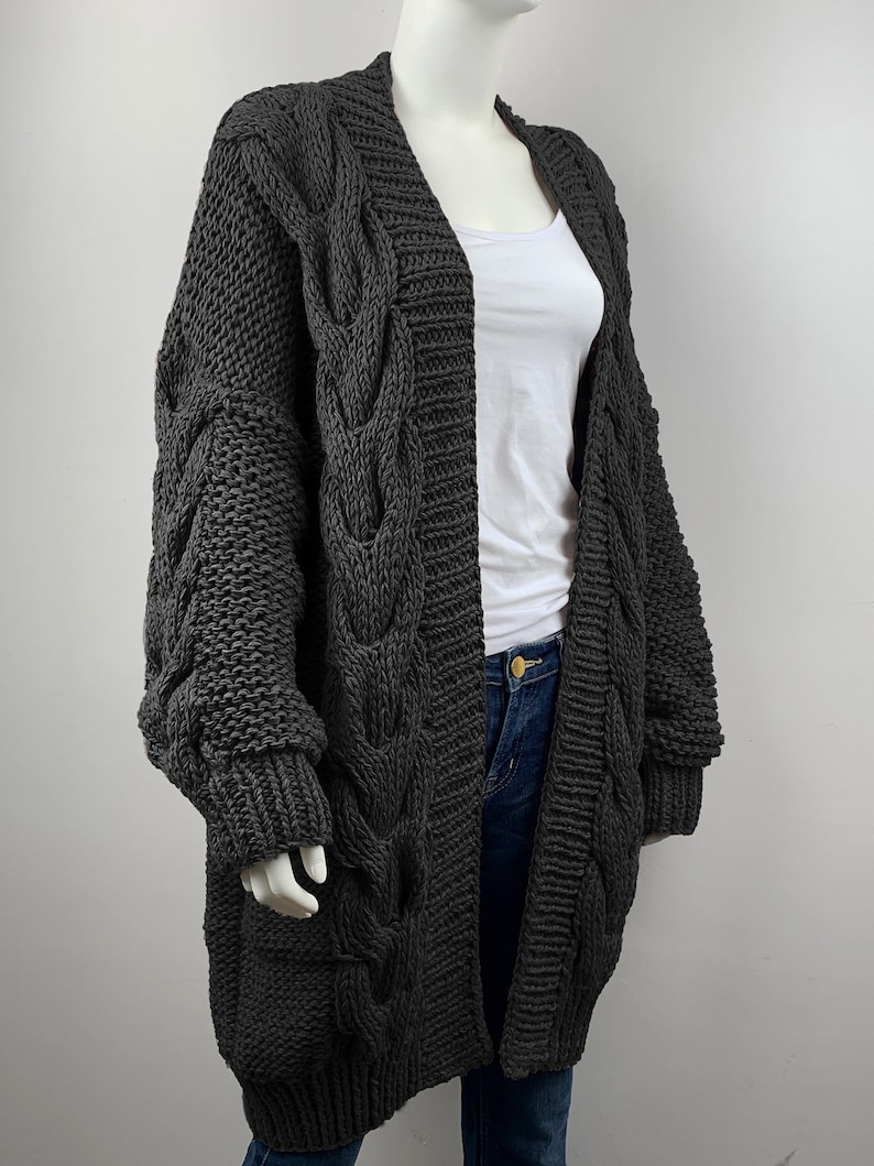 May include: A long, gray cable knit cardigan with a V-neck and long sleeves. The cardigan is made of a chunky yarn and has a relaxed fit.