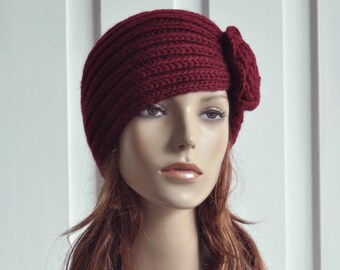 May include: A burgundy knitted headband with a large flower accent. The headband is worn on a mannequin with long brown hair.