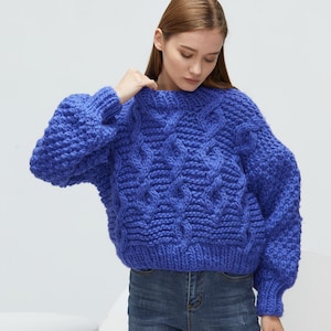Puede incluir: Un suéter azul de punto grueso con un patrón de punto trenzado. El suéter tiene un corte holgado y mangas largas.