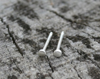 May include: Two small, silver-colored stud earrings with a flat, circular design. The earrings are set against a weathered, gray wooden surface, creating a rustic contrast. The studs are simple and minimalist, suitable for everyday wear.