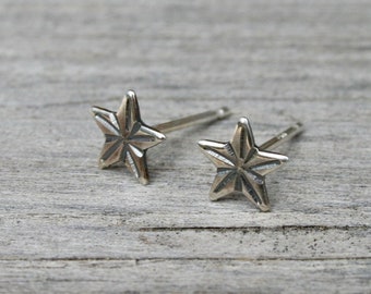 May include: Two silver-colored star-shaped stud earrings. Each earring has a five-pointed star design with detailed facets. The earrings are displayed on a weathered, gray wooden surface, creating a rustic backdrop for the jewelry.