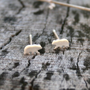 May include: A pair of small, silver-colored bear stud earrings. The earrings are in the shape of a bear silhouette and are set against a textured, gray wooden surface. The bear studs are a simple, nature-inspired accessory.
