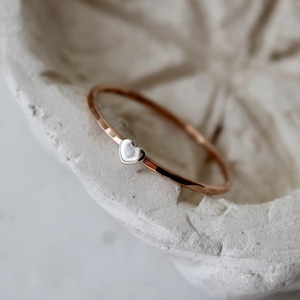 May include: A rose gold bangle bracelet with a small silver heart charm. The bracelet is resting in a white, textured, decorative bowl. The bangle has a hammered texture.