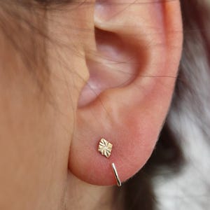 May include: A close-up of an ear with two earrings. One is a small, textured, gold-colored stud in a diamond shape. The other is a small, silver-colored hoop earring.
