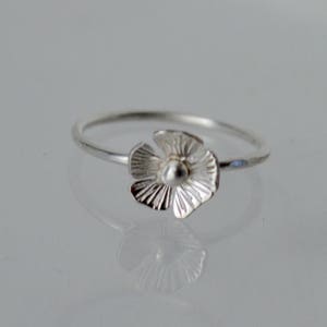 May include: A silver ring featuring a small, five-petaled flower charm. The flower has textured, radiating lines on each petal and a smooth, rounded center. The band of the ring is thin, polished, and circular, reflecting light on its metallic surface.
