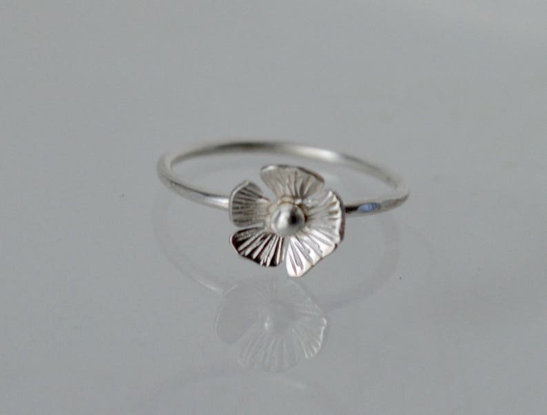 May include: A silver ring featuring a small, five-petaled flower charm. The flower has textured, radiating lines on each petal and a smooth, rounded center. The band of the ring is thin, polished, and circular, reflecting light on its metallic surface.
