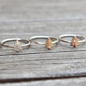 May include: Three delicate rings with a tree-shaped charm. The rings have a thin band and a small, textured tree design. The rings are made of silver and gold-colored metal. The rings are displayed on a wooden surface.
