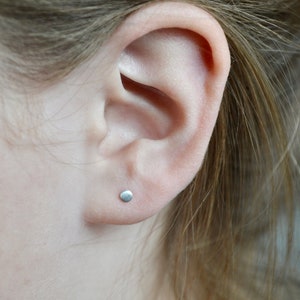 May include: A close-up view of a person's ear with a small, round, silver stud earring. The earring is centered on the earlobe. The person's hair is light brown and partially obscures the ear.
