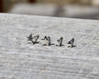 May include: Four silver star-shaped stud earrings of varying sizes are arranged on a wooden surface. The earrings are small and shiny, with a simple, five-pointed star design. The studs are likely made of metal.