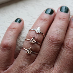 May include: Three silver rings, each featuring a small tree design, are displayed on a hand with dark green nail polish. The rings are simple and delicate, showcasing a nature-inspired theme.