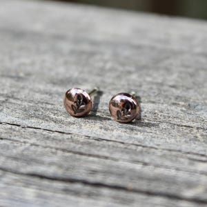 May include: A pair of small, round, metallic copper-toned stud earrings. Each earring features a polished, domed surface with a small, dark, engraved leaf motif centered on the front. The earrings have a simple, circular shape and a reflective finish.