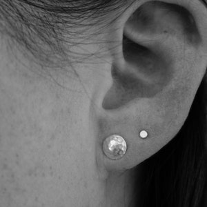 May include: A close-up of a person's ear with two small earrings. The earring on the lower lobe is a hammered silver disc, and the earring on the upper lobe is a small silver stud.
