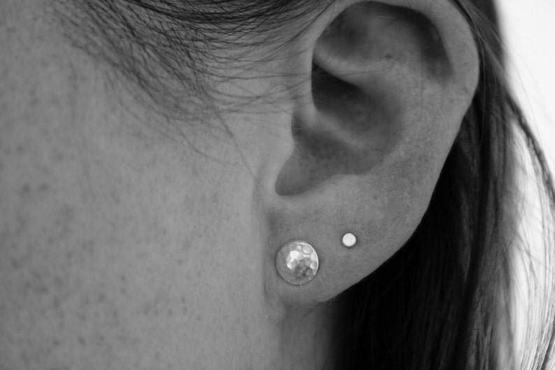 May include: A close-up of a person's ear with two small earrings. The earring on the lower lobe is a hammered silver disc, and the earring on the upper lobe is a small silver stud.