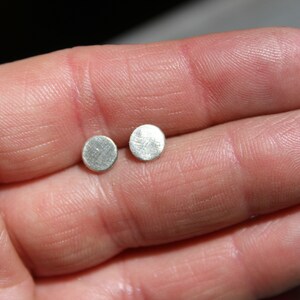 Peut inclure: Deux petites boucles d'oreilles rondes de couleur argentée. Chaque boucle d'oreille présente une surface texturée et un design simple et minimaliste. Les boucles d'oreilles sont présentées sur un fond neutre, soulignant leur forme et leur éclat métallique. Bijou classique.