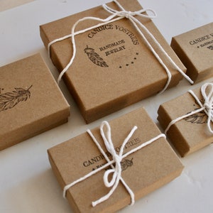 May include: Four brown cardboard boxes with white string tied around them. Each box has a different design with a feather and the text "Candice Vosreis Handmade Jewelry".