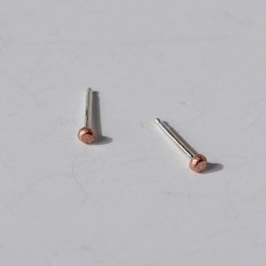 May include: Two small stud earrings. Each earring has a silver post with a copper-colored, rounded end. The earrings are displayed on a white surface, with one earring slightly angled and the other upright.