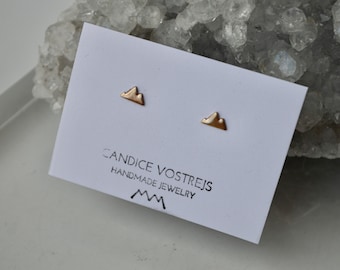Puede incluir: Un par de pendientes de oro en forma de montaña sobre una tarjeta blanca con el texto "Candice Vostrejs Handmade Jewelry" y un logotipo de montaña estilizado.