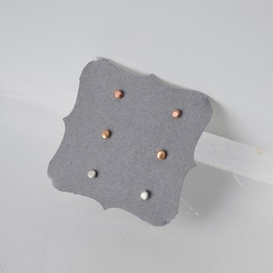 May include: A set of three small, round stud earrings in silver, gold, and rose gold. The earrings are displayed on a gray card.