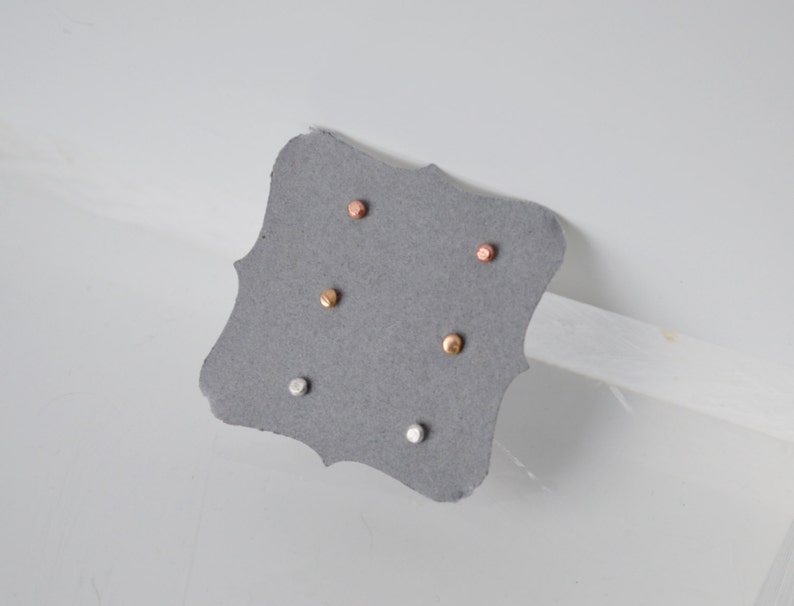 May include: A set of three small, round stud earrings in silver, gold, and rose gold. The earrings are displayed on a gray card.