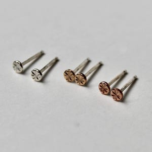 May include: A collection of stud earrings in silver and rose gold. Each earring features a small, round, textured design on the front. The earrings are displayed on a white surface, showcasing their delicate details and metallic shine.