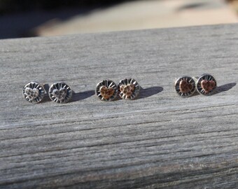 May include: Three pairs of circular stud earrings featuring a central heart shape. The first pair is silver with a hammered texture, the second pair has a copper heart on a silver base, and the third pair features copper hearts on silver discs.